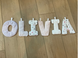 A set of six baby foot-shaped footies arranged vertically on a light brown wooden floor, spelling out the word 'LOVE'. The footies are made of soft, textured fabric in pastel shades of pink, white, and light blue with delicate floral prints.