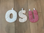 Three fabric baby neck pillows in different patterns (solid cream, beige with white polka dots, and red gingham) arranged vertically on a light brown wooden floor.