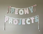 Custom fabric letter banner spelling 'PEONY PROJECTS' in pastel pink, white, and light blue-green, hung on a gray wall with pink ribbon ties. The image has a cream border with the text 'Custom Banners' at the top.