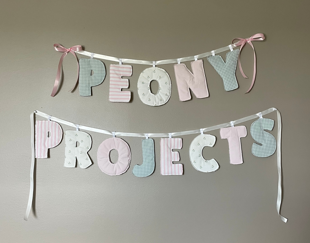 Custom fabric letter banner spelling 'PEONY PROJECTS' in pastel pink, white, and light blue-green, hung on a gray wall with pink ribbon ties. The image has a cream border with the text 'Custom Banners' at the top.
