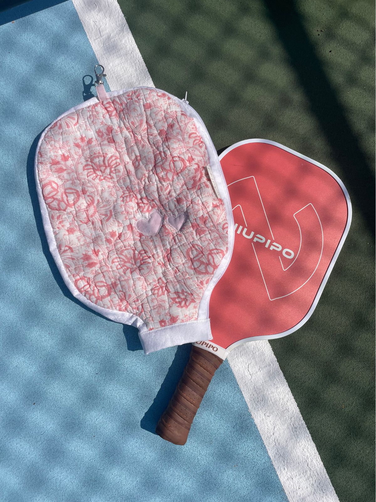 A close-up of a paddle racket with a red head featuring the brand name 'NUPISO' in white, and a quilted white cover with a pink floral pattern, resting on a blue and green court surface.