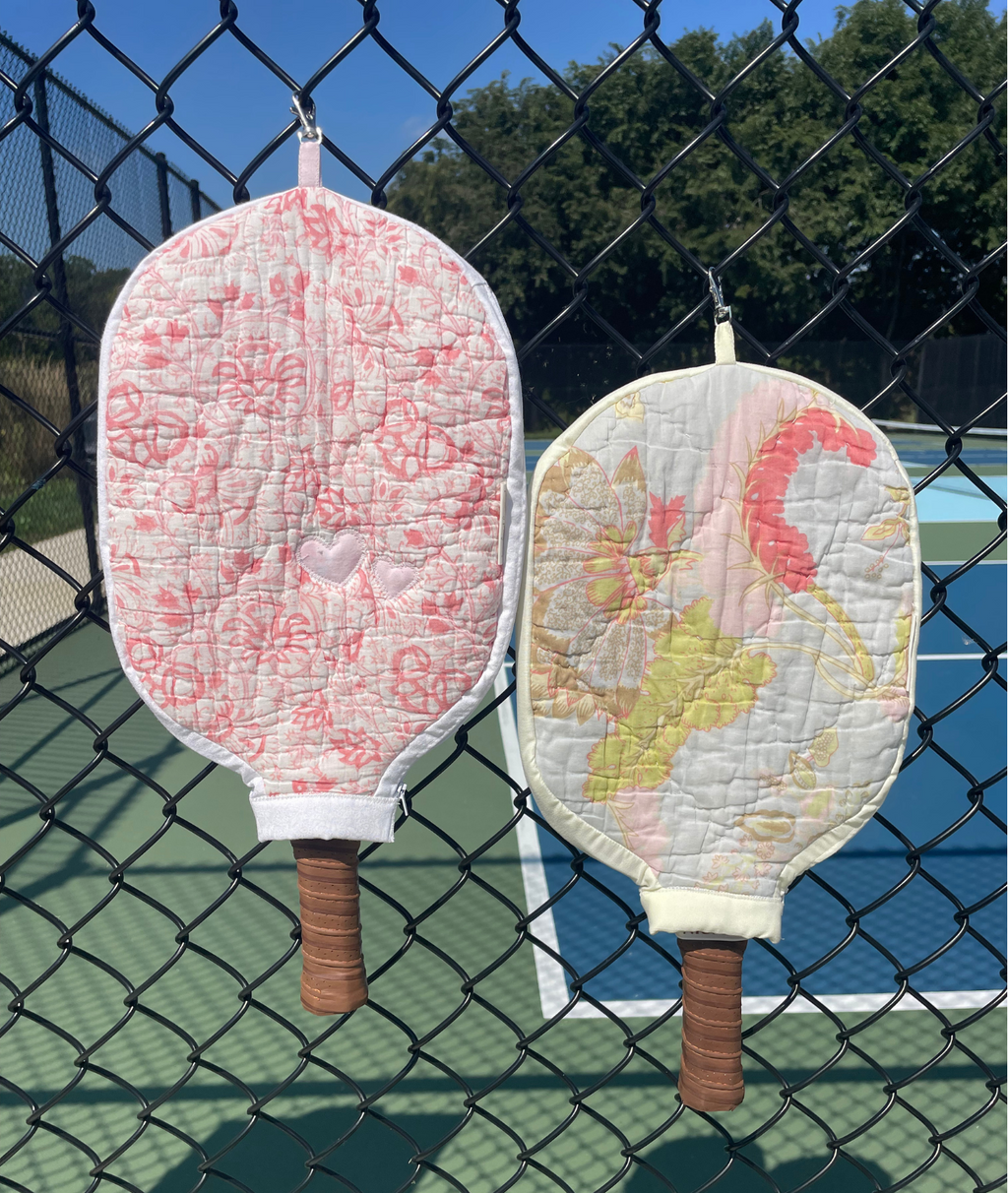 Two quilted pickleball paddles with fabric covers hang on a black chain-link fence in front of a blue tennis court. One cover is light pink with a darker pink floral pattern, and the other is light yellow with a pink flamingo design.