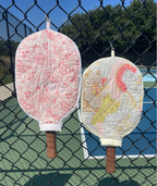 Two quilted pickleball paddles with fabric covers hang on a black chain-link fence in front of a blue tennis court. One cover is light pink with a darker pink floral pattern, and the other is light yellow with a pink flamingo design.