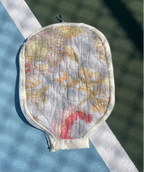 A quilted tennis ball bag with a floral pattern, placed on a blue and green tennis court near a white boundary line.