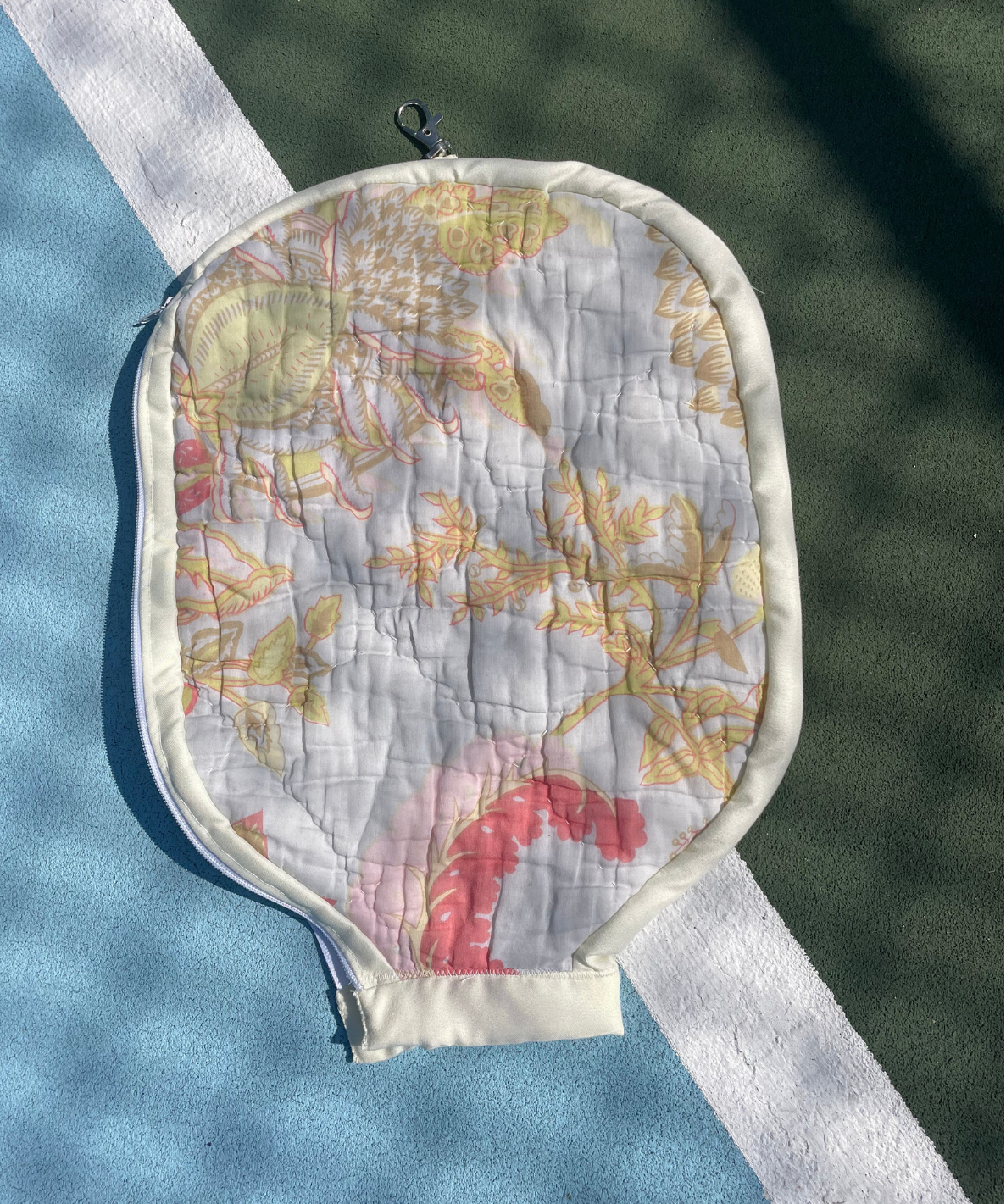 A quilted tennis ball bag with a floral pattern, placed on a blue and green tennis court near a white boundary line.