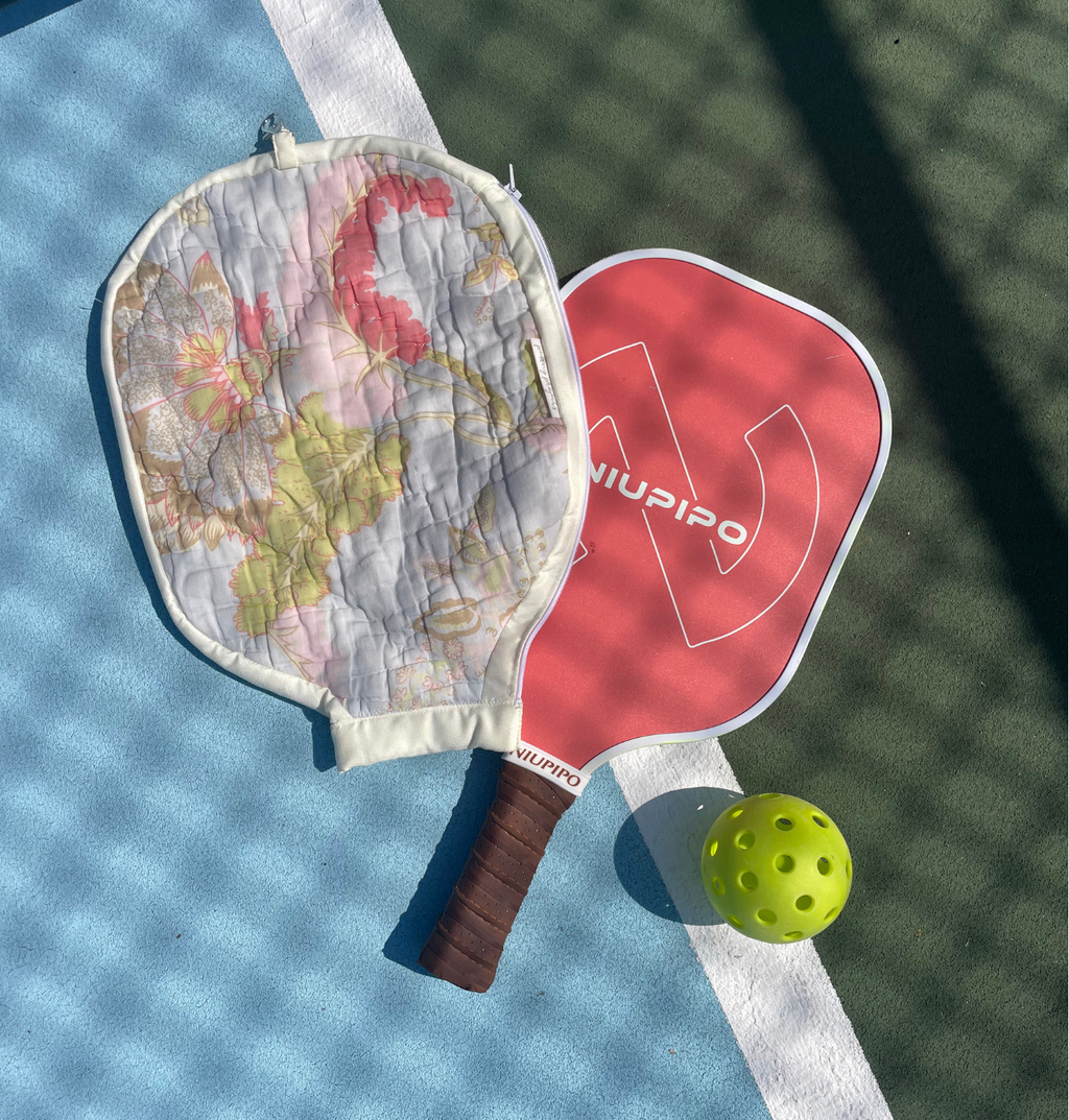Two pickleball paddles and a yellow ball arranged on a blue and green pickleball court. One paddle has a floral quilted cover, and the other is red with the brand name 'Niupipo' visible.