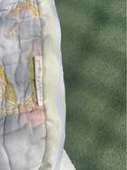 A close-up of a quilted fabric item with a floral print in pale blue, pink, and yellow, showing a white fabric tag with handwritten text, next to a green textured surface.