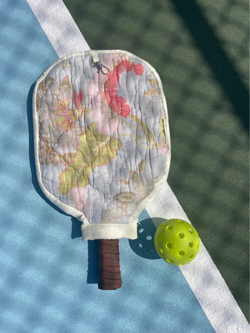 A quilted pickleball paddle cover with a vintage map print and a brown grip, shown on a tennis court alongside a yellow pickleball.
