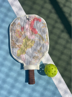 A quilted pickleball paddle cover with a vintage map print and a brown grip, shown on a tennis court alongside a yellow pickleball.