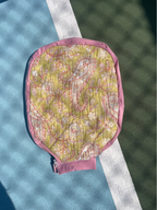 A quilted paddle-style tennis cover with a pink paisley pattern on a yellow-green background, featuring a solid pink border and a pink zipper tab, laid flat on a blue and green tennis court.