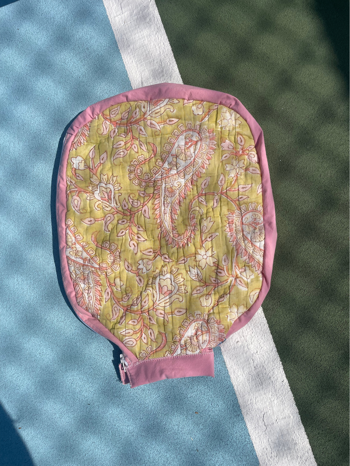 A quilted paddle-style tennis cover with a pink paisley pattern on a yellow-green background, featuring a solid pink border and a pink zipper tab, laid flat on a blue and green tennis court.