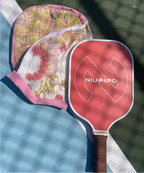 A red Odenin pickleball paddle and a matching pink and green floral quilted pad laid out on a blue and green pickleball court.