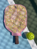 A quilted pickleball paddle cover with a pink paisley pattern and brown handle grip, shown on a blue and green pickleball court next to a yellow pickleball.