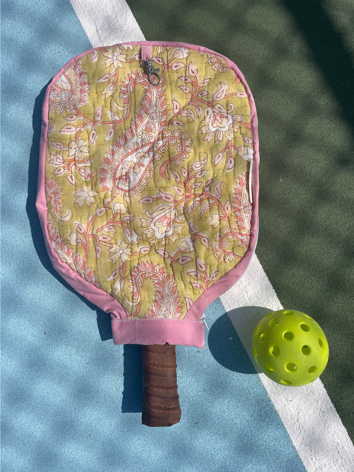 A quilted pickleball paddle cover with a pink paisley pattern and brown handle grip, shown on a blue and green pickleball court next to a yellow pickleball.
