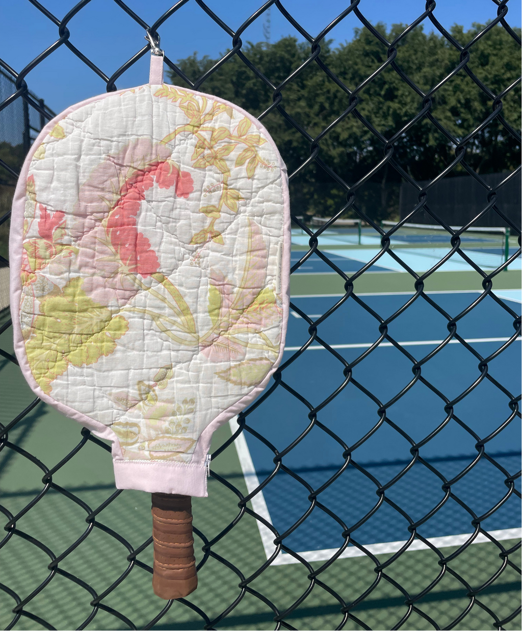 A quilted pickleball paddle cover with a pink, yellow, and white floral pattern, hanging on a black chain-link fence in front of a blue pickleball court.