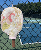 A quilted pickleball paddle cover with a pink, yellow, and white floral pattern, hanging on a black chain-link fence in front of a blue pickleball court.