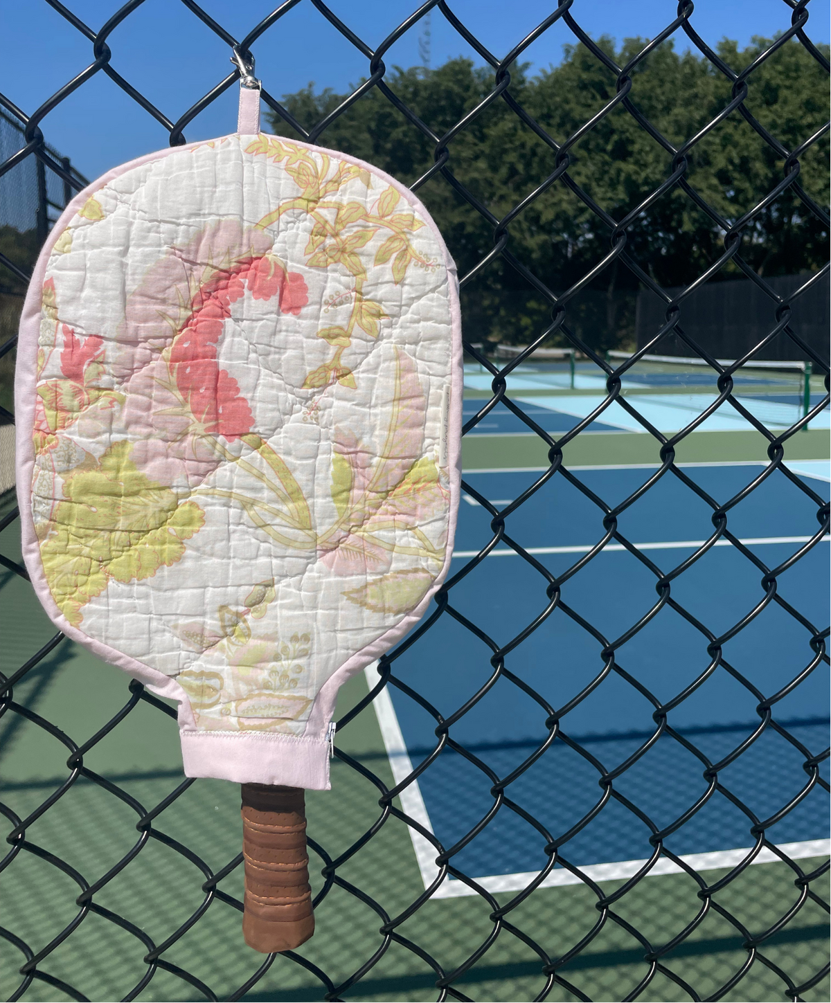 A quilted pickleball paddle cover with a pink, yellow, and white floral pattern, hanging on a black chain-link fence in front of a blue pickleball court.