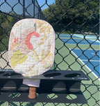 A quilted pickleball paddle pad with a pink, yellow, and white floral pattern, mounted on a black rack on a chain-link fence at an outdoor pickleball court.