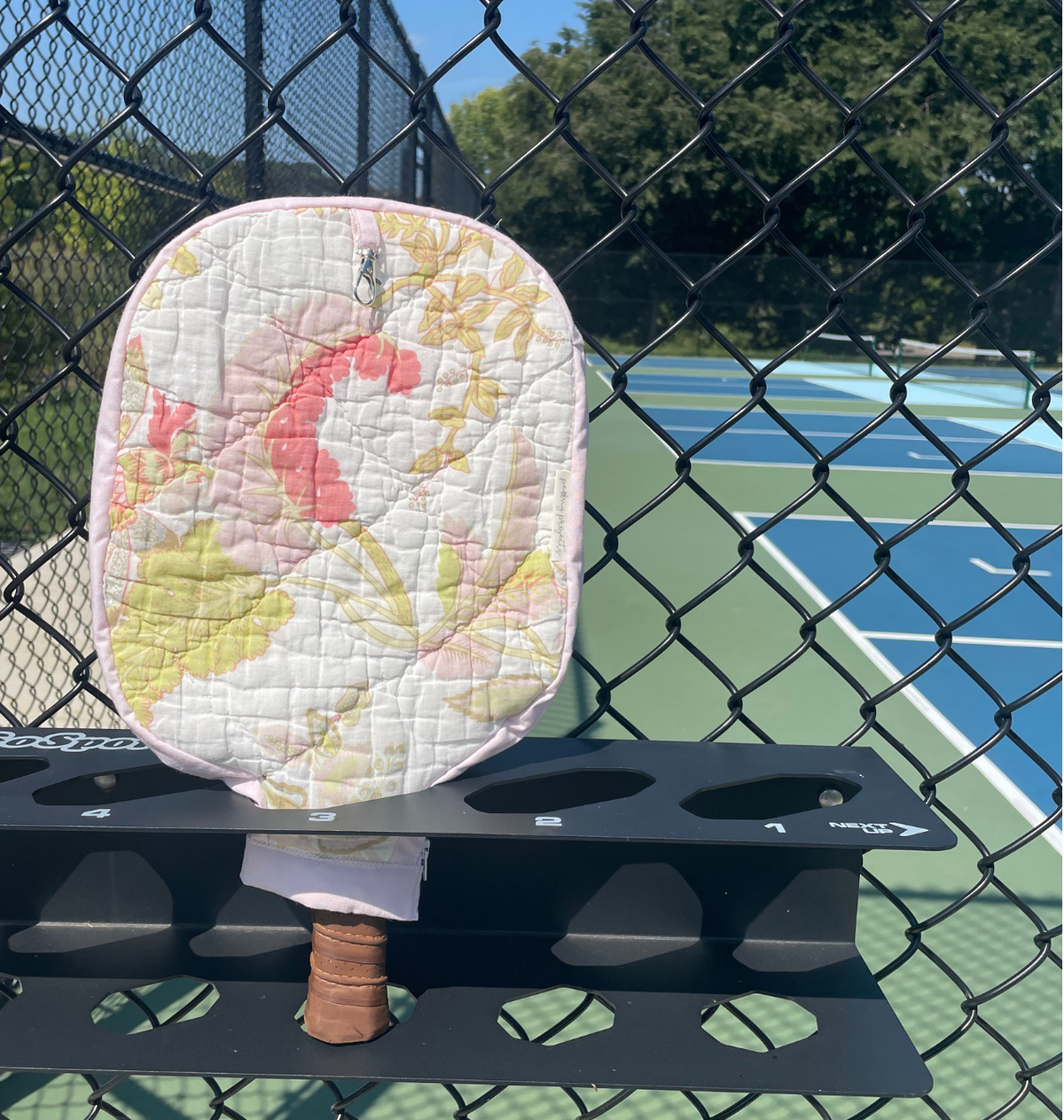 A quilted pickleball paddle pad with a pink, yellow, and white floral pattern, mounted on a black rack on a chain-link fence at an outdoor pickleball court.