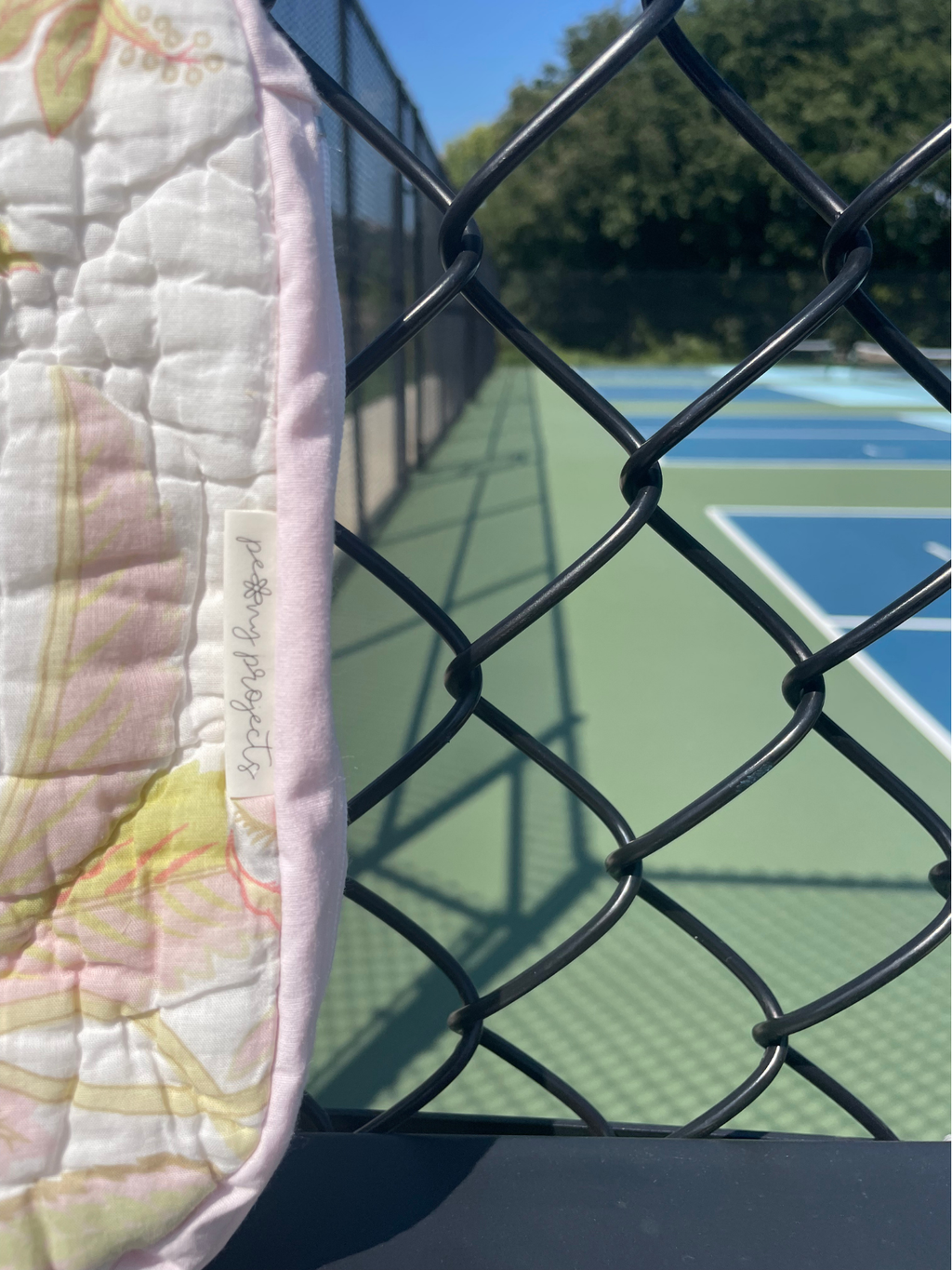 A close-up of a quilted fabric panel with a pastel floral pattern and a visible handwritten tag reading 'Personalized', positioned behind a black chain-link fence with a tennis court in the background.
