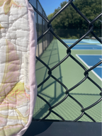 A close-up of a quilted fabric panel with a pastel floral pattern and a visible handwritten tag reading 'Personalized', positioned behind a black chain-link fence with a tennis court in the background.