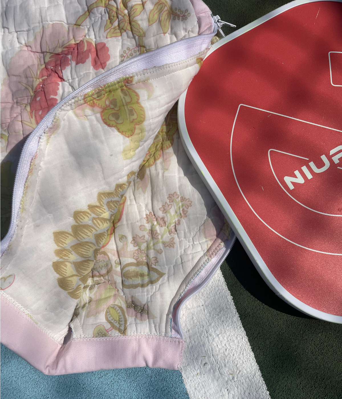 A close-up of a quilted athletic support garment with a floral pattern in soft pink, green, and beige tones, shown next to a red and white running shoe on a textured green and blue surface.