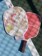 A pickleball paddle with a pink and white graphic face and a brown wrapped handle, placed on a blue and green tennis court alongside a matching quilted paddle cover featuring a floral print.
