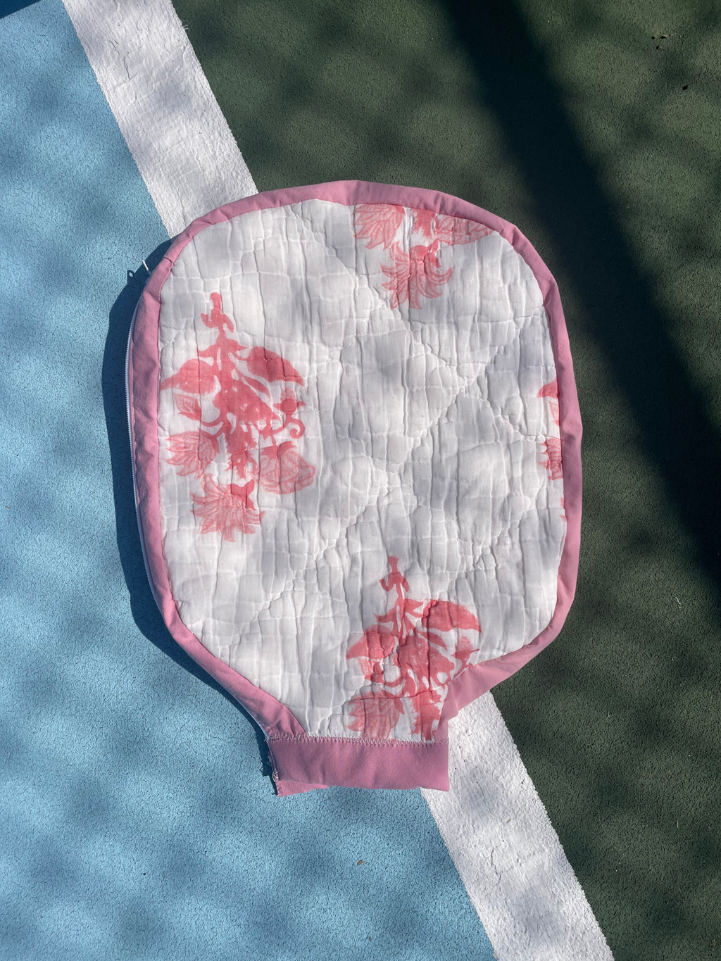 A quilted paddle-shaped tennis ball grip cover with a pink floral pattern on a white background, bordered in pink, laid on a blue and green tennis court.