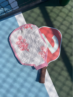 A quilted pickleball paddle cover with a pink floral pattern and a pink border, shown on a blue and green pickleball court next to a paddle with a red and white logo.