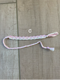 A handmade braided friendship bracelet with a pink and white pattern, featuring a tassel and a small white bead, laid flat on a light brown wood-grain surface.