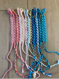 Six braided friendship bracelets in pink, cream, yellow, blue, and teal, arranged vertically on a light brown fabric background.