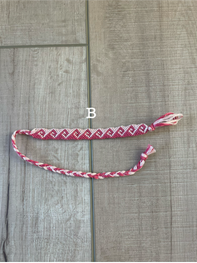 A handmade braided friendship bracelet with a red and white pattern, laid out on a gray wood plank floor. A small white label with the letter 'B' is placed near the center of the bracelet.