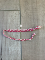 A handmade braided friendship bracelet with a red and white pattern, laid out on a gray wood plank floor. A small white label with the letter 'B' is placed near the center of the bracelet.